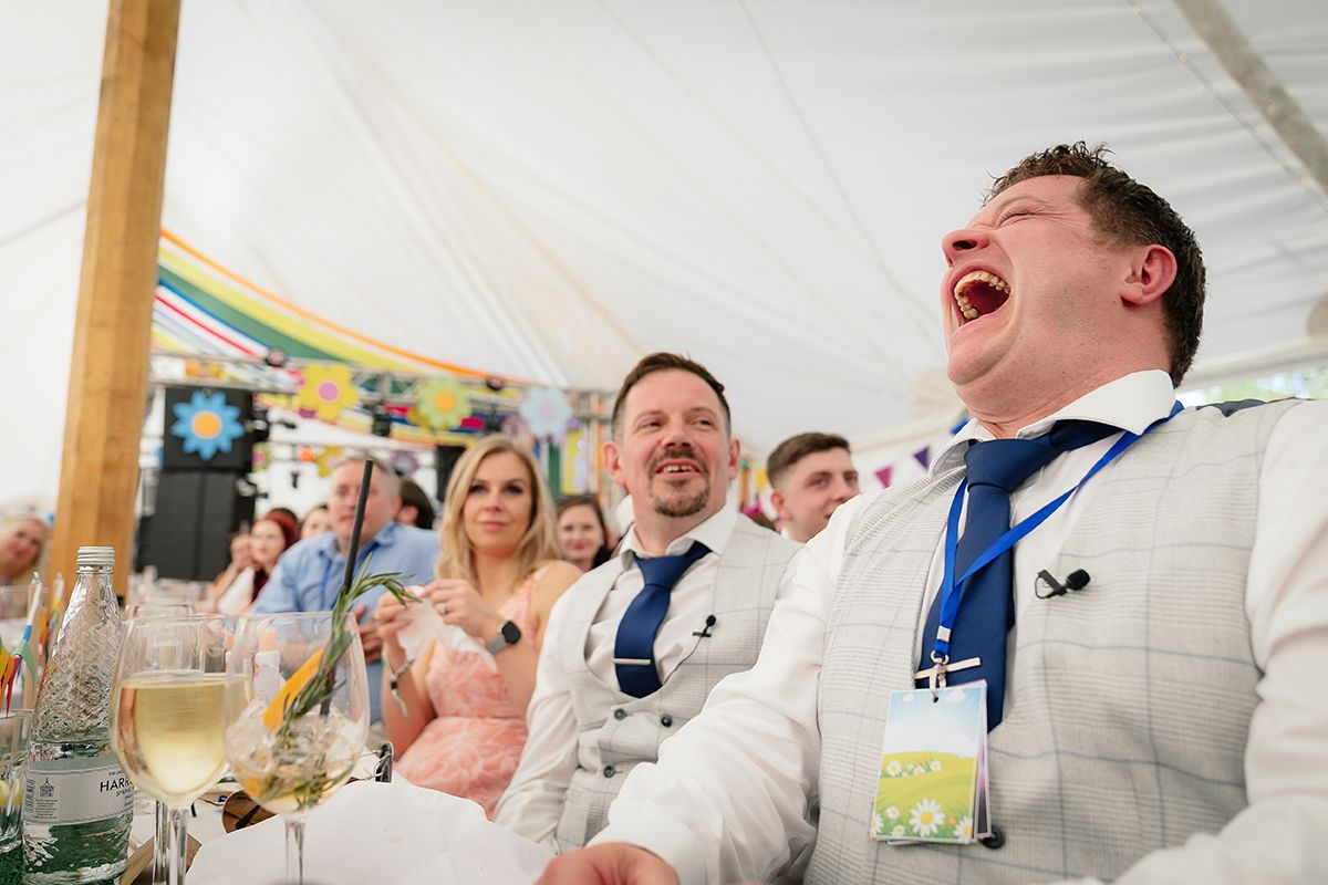 Wedding party members laugh and celebrate during speeches at an indoor reception.