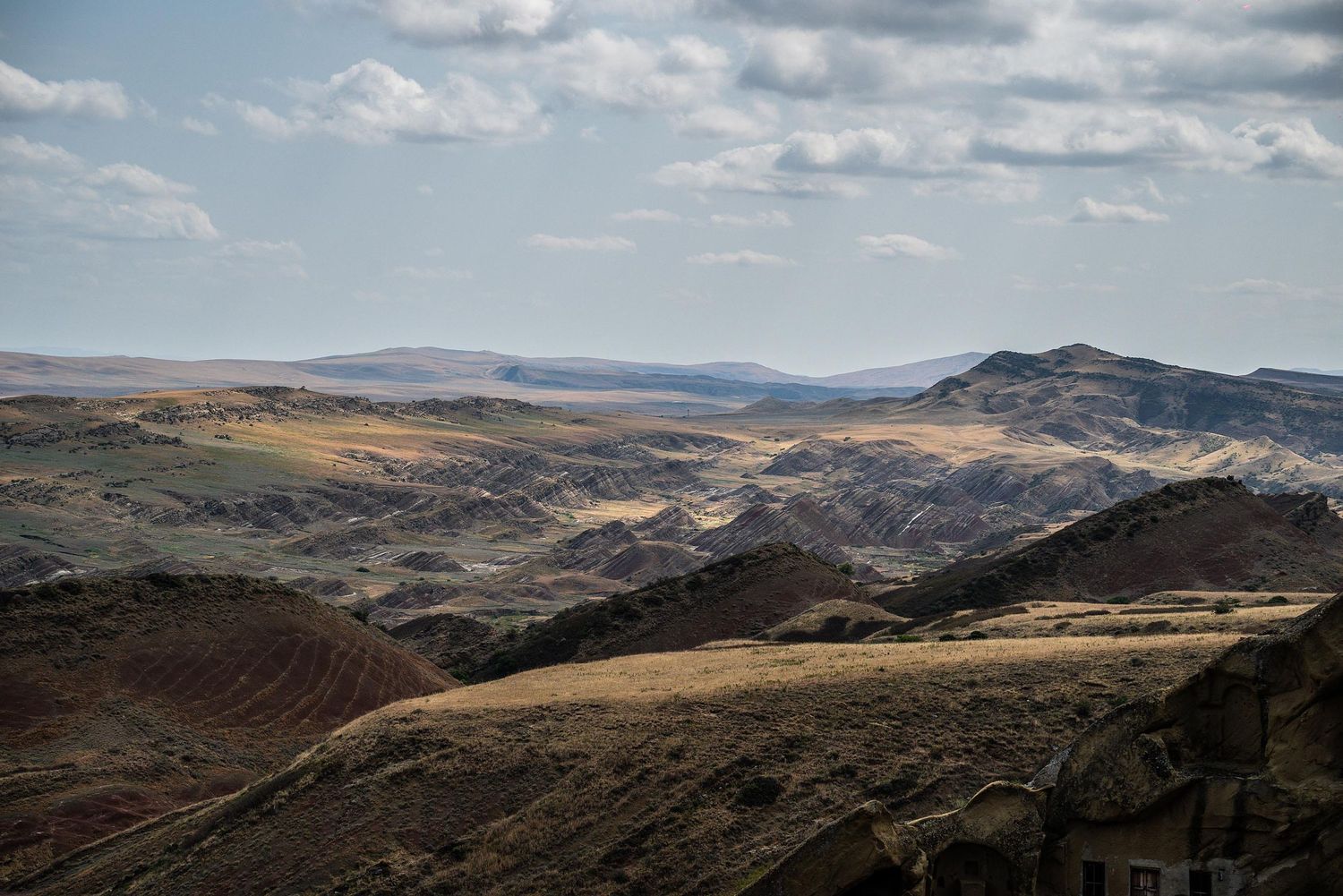Panoramic view of rolling hills and mountain ranges under a cloudy blue sky in a remote wilderness landscape.