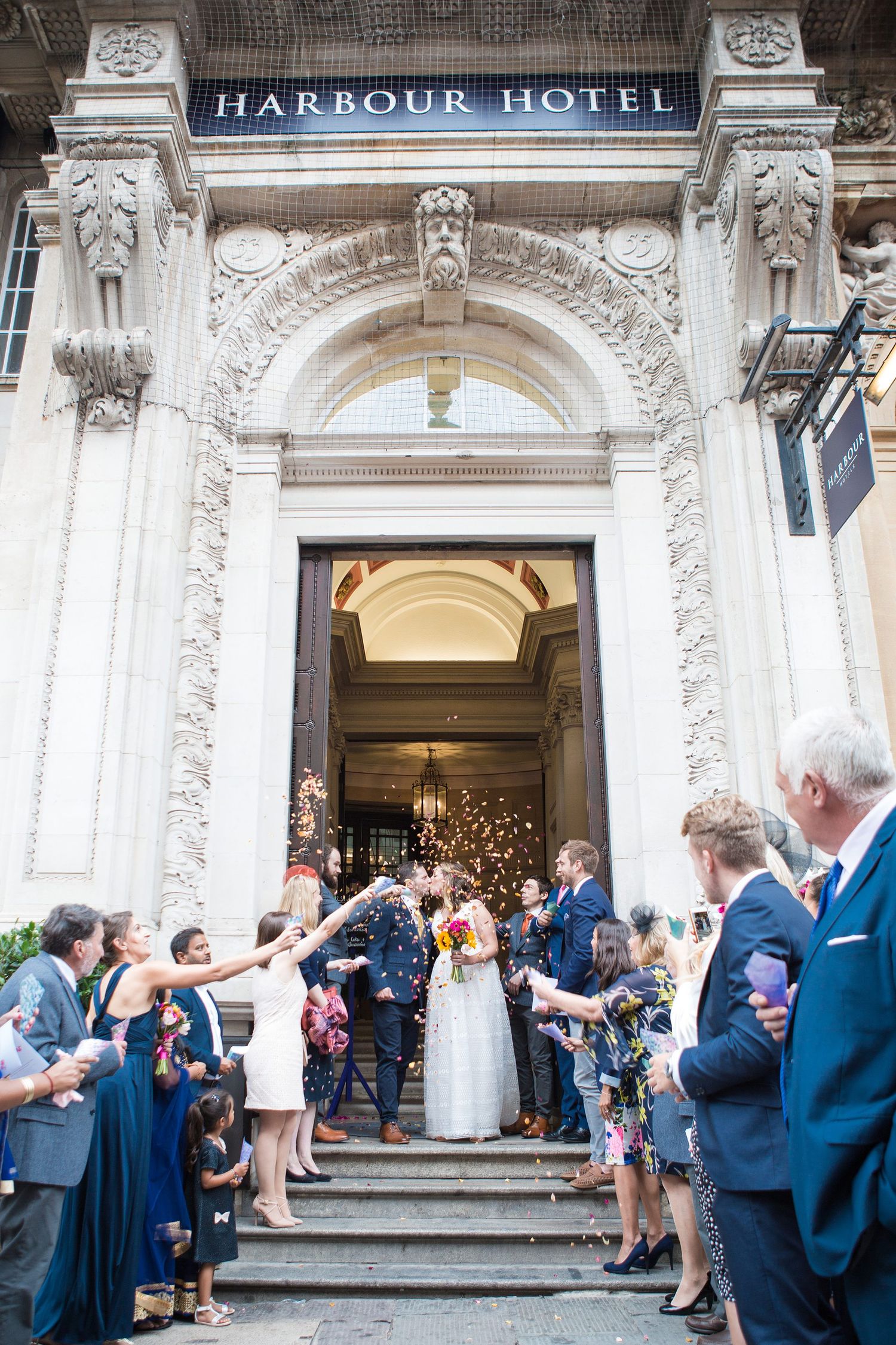 Wedding guests throw confetti as newlyweds exit the grand entrance of the Harbour Hotel with ornate stone architecture.