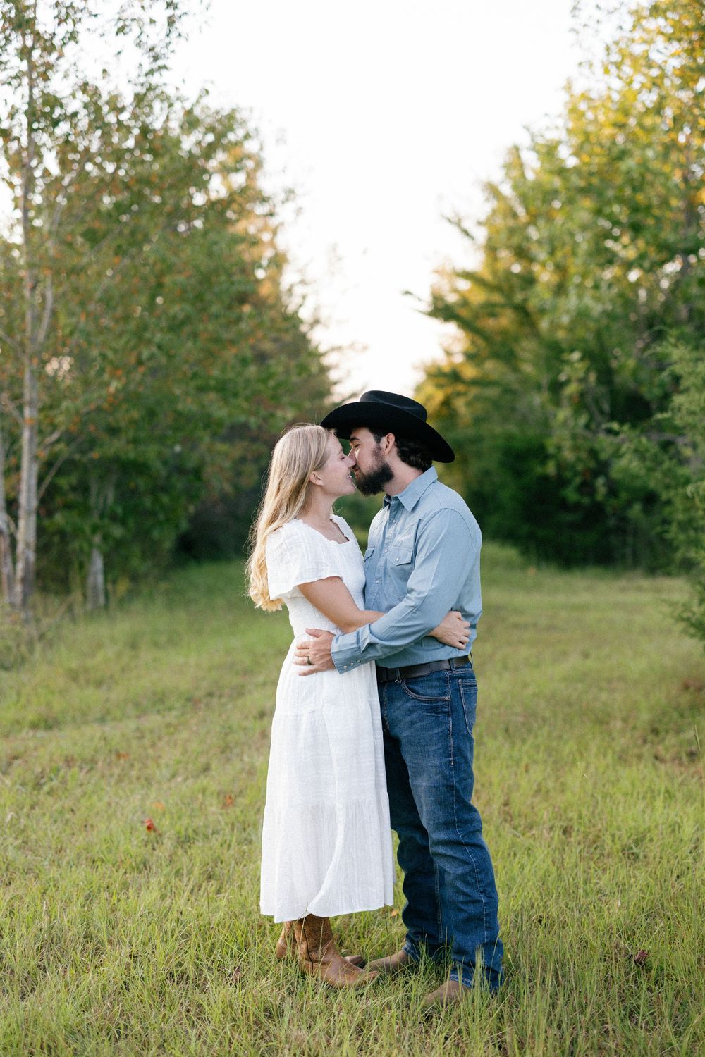 A couple embraces in a sunlit meadow, with her in a white dress and him wearing a cowboy hat and jeans.