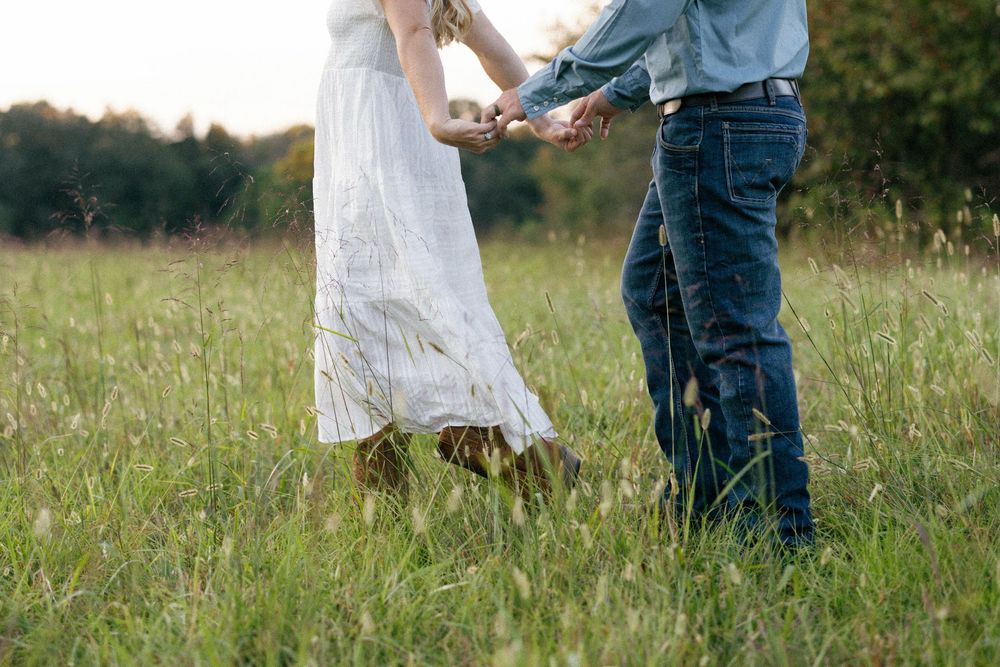 A couple in casual attire embraces while standing together in a sunlit grassy meadow during golden hour.