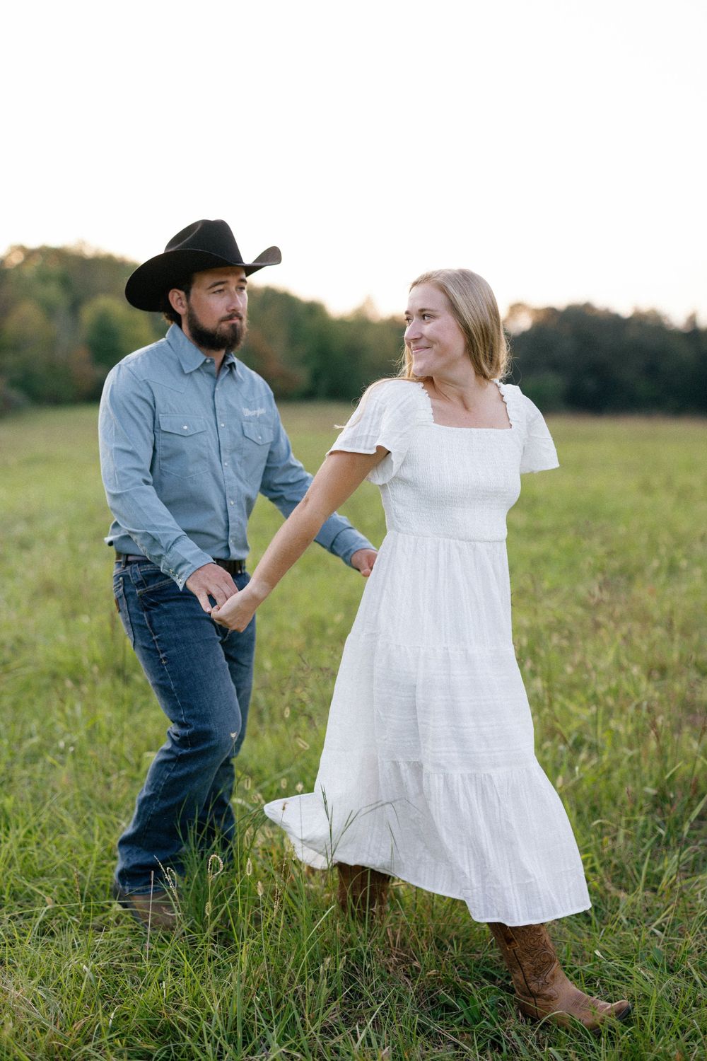 A couple in western attire playfully runs through a grassy field at sunset.