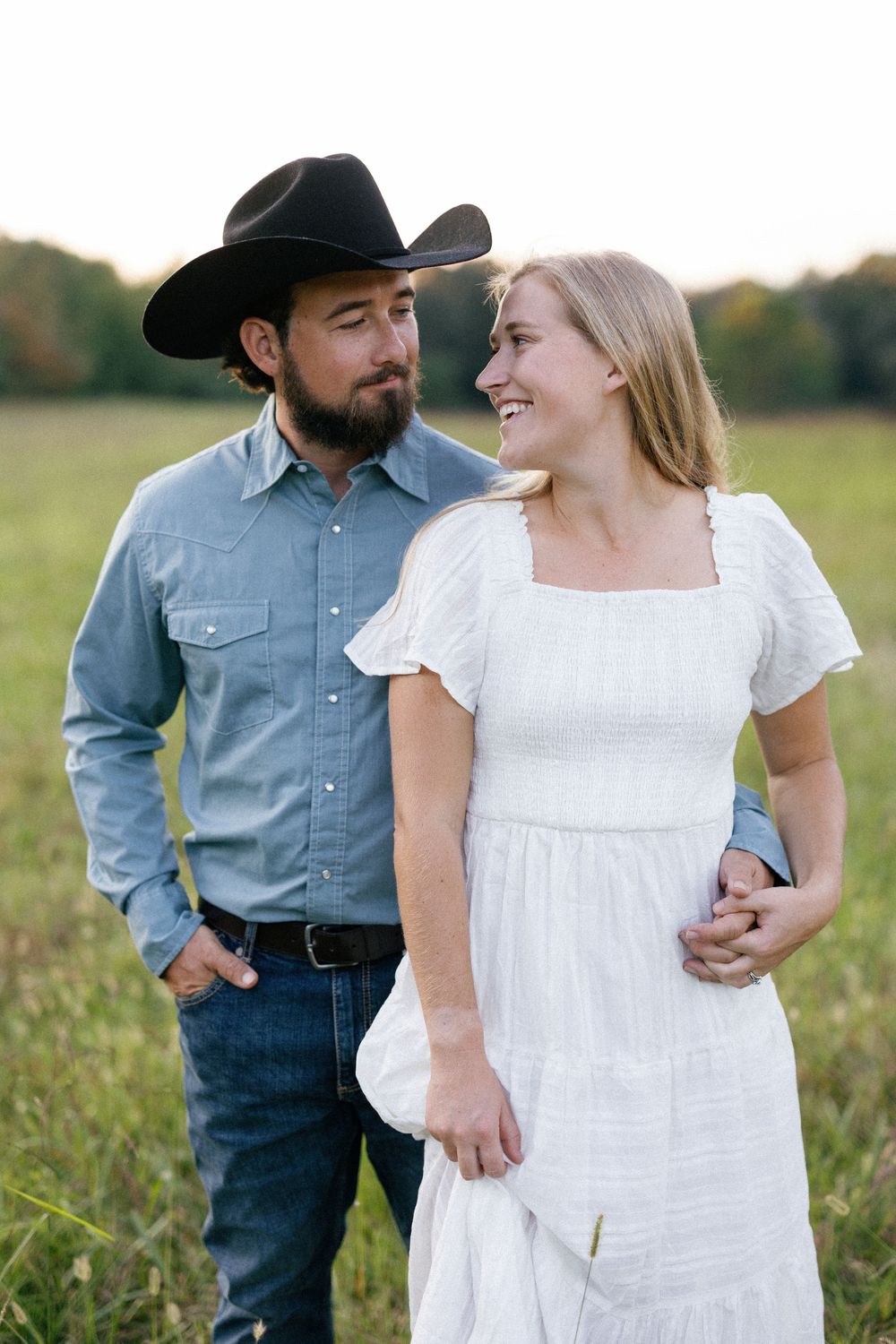 A couple stands together in a field at sunset, with one wearing a cowboy hat and denim shirt and the other in a white dress.