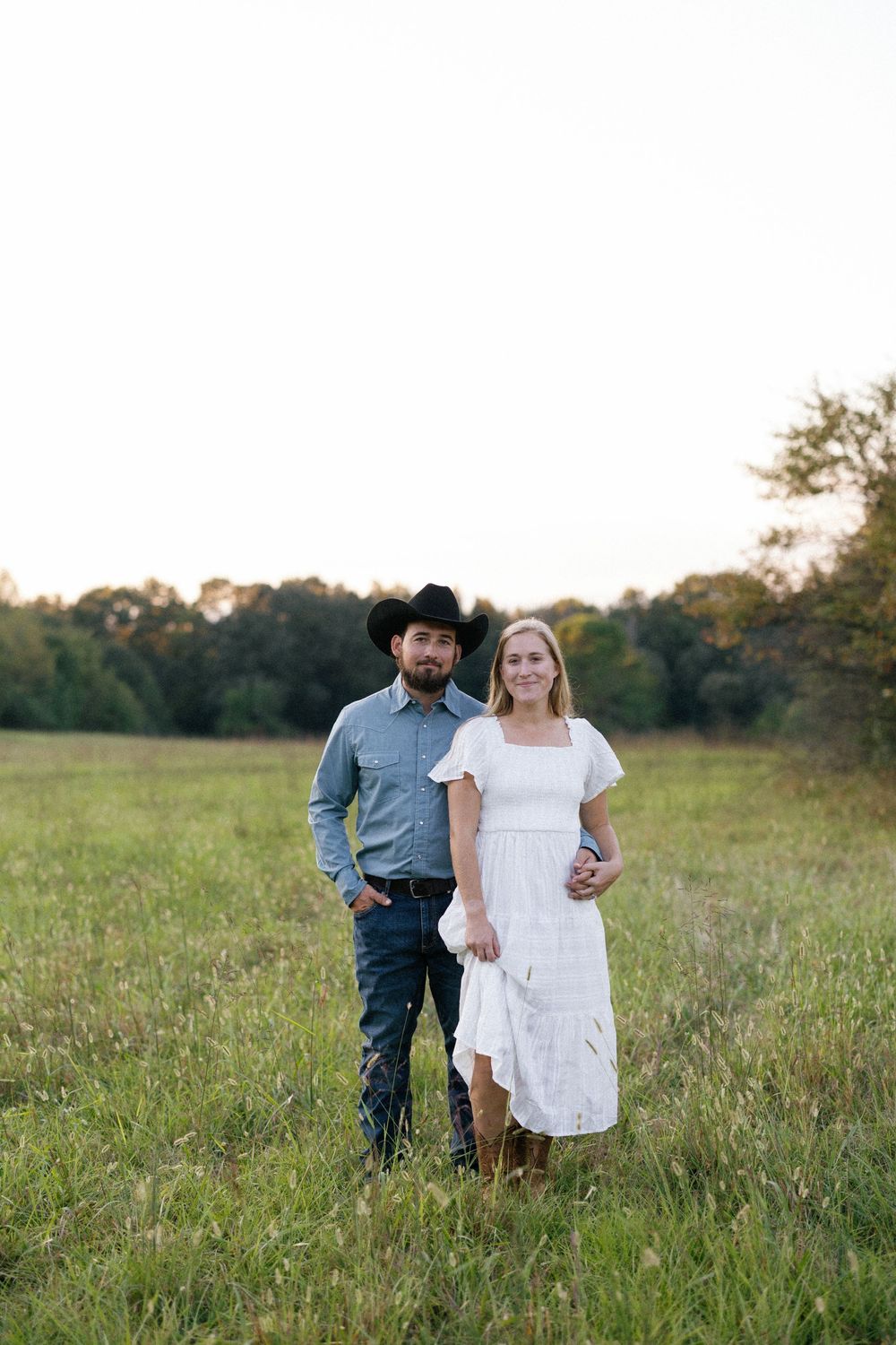A couple in western attire poses together in a sunny meadow during an engagement photoshoot.