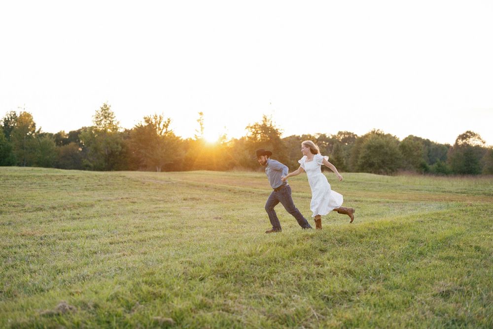 Two people dance joyfully in a grassy field at sunset with golden light glowing behind them.