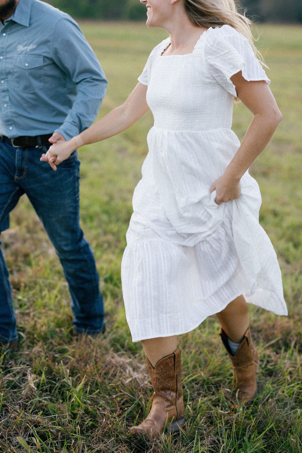 A person in a flowing white dress and cowboy boots stands in a grassy field next to someone wearing jeans.