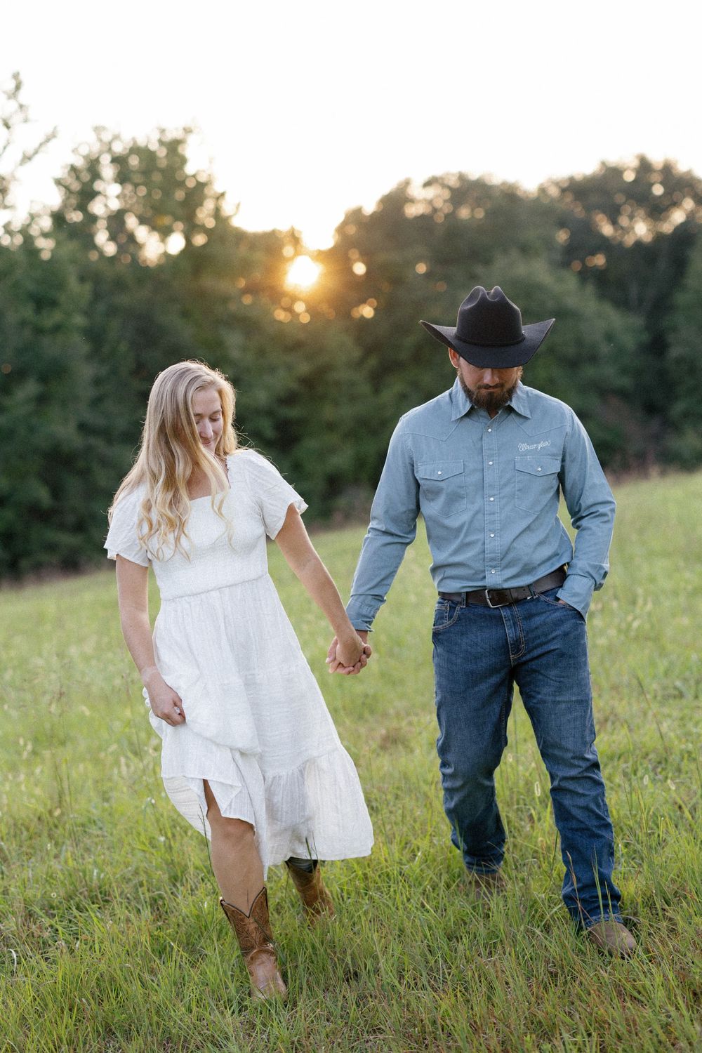 A couple walks hand in hand through a sunlit meadow at golden hour, wearing casual western attire.