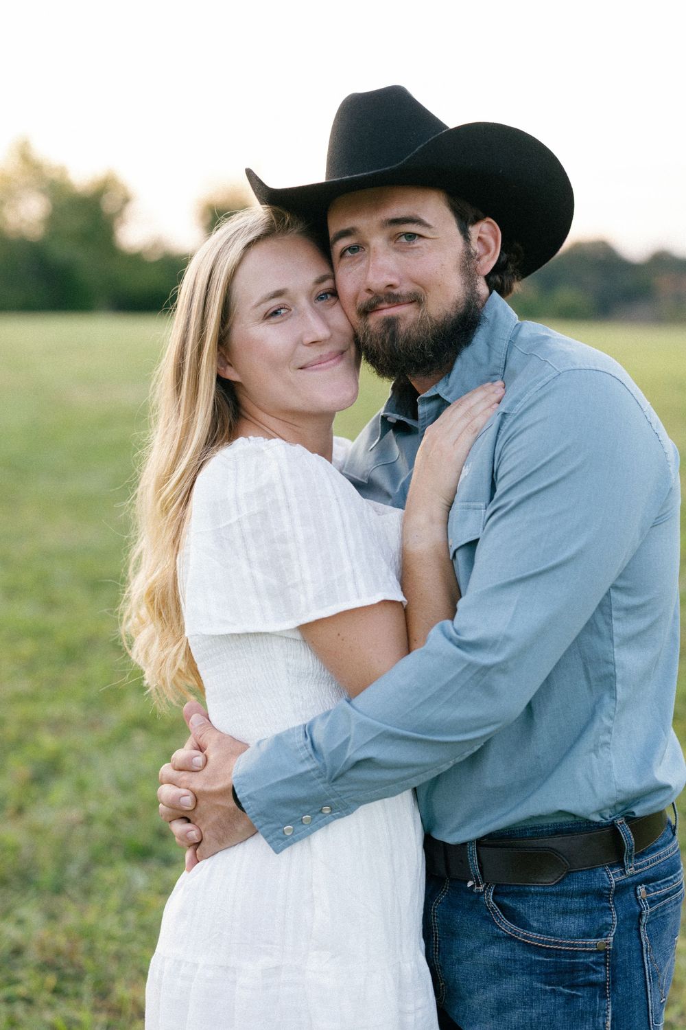 A couple in western attire embraces in a sunlit field during a romantic photoshoot.