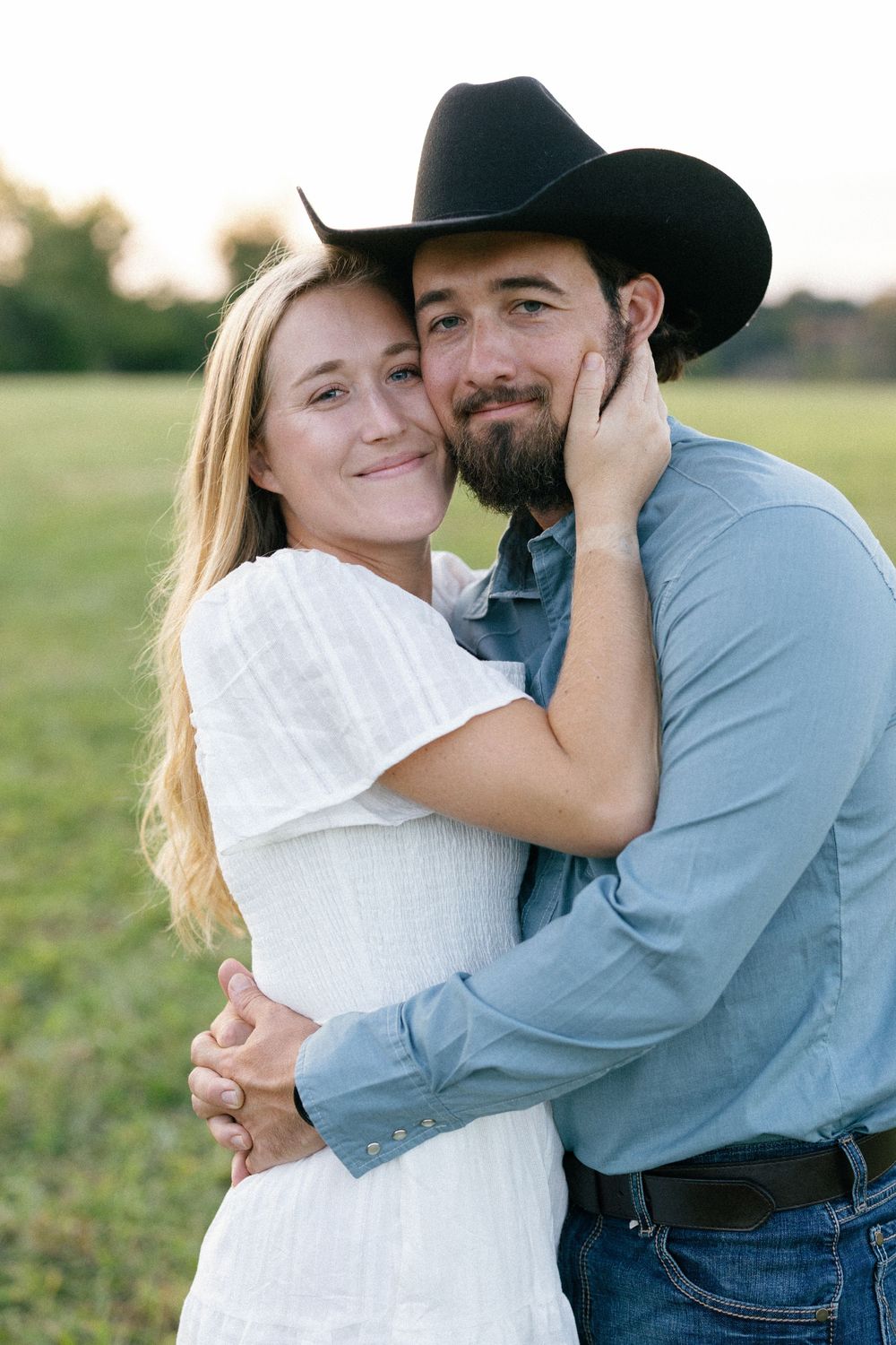 A couple in western attire shares tender embraces in a sunny field during a romantic outdoor photoshoot.