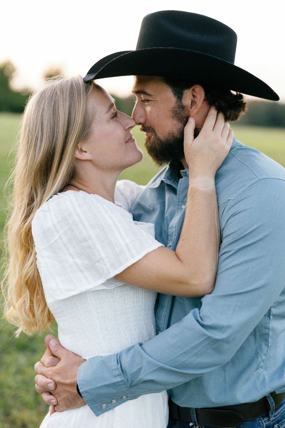 A romantic couple embraces and shares a tender kiss outdoors, with the person wearing a cowboy hat and blue shirt.