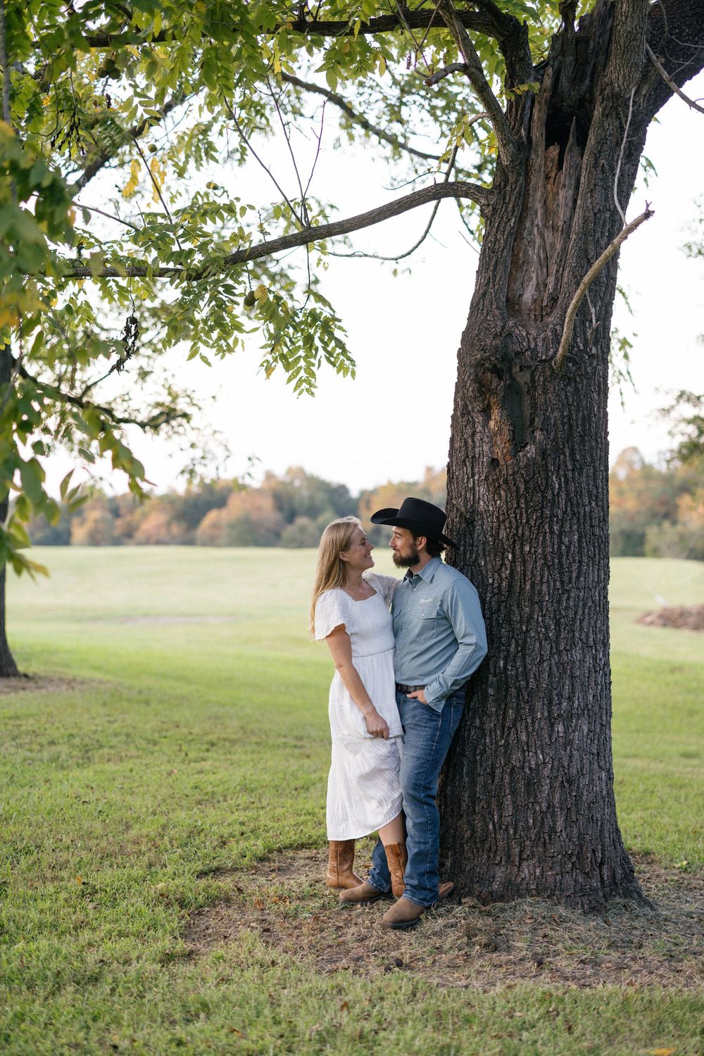 A couple shares a romantic moment under a large tree in a grassy field during autumn.