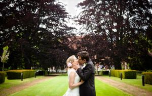 A couple embraces in a romantic moment along a tree-lined garden path with manicured hedges and green lawns.