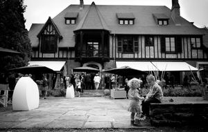 Vintage black and white photograph of people gathering outside a Tudor-style house with awnings and outdoor seating area.