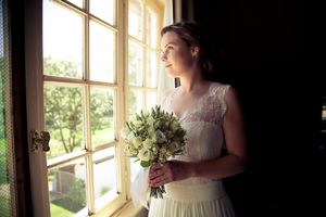 A bride in a white lace gown holds a bouquet of white flowers while standing by a sunlit window.