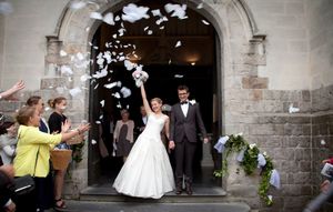 Wedding guests throw white rose petals as newlyweds exit stone church doorway during celebration.