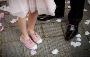 Pink rose petals scattered on a concrete walkway with glimpses of formal shoes and dress hems.