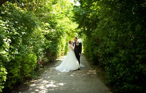 A couple in formal wedding attire walks down a sunlit garden path surrounded by lush green foliage.