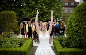 A joyful bride raises her arms in celebration while standing in a formal garden with guests in the background.