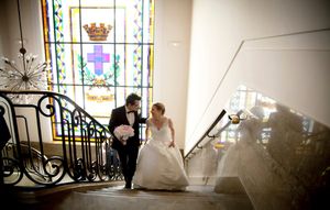 A couple in wedding attire ascends an elegant staircase with ornate iron railings and stained glass windows.