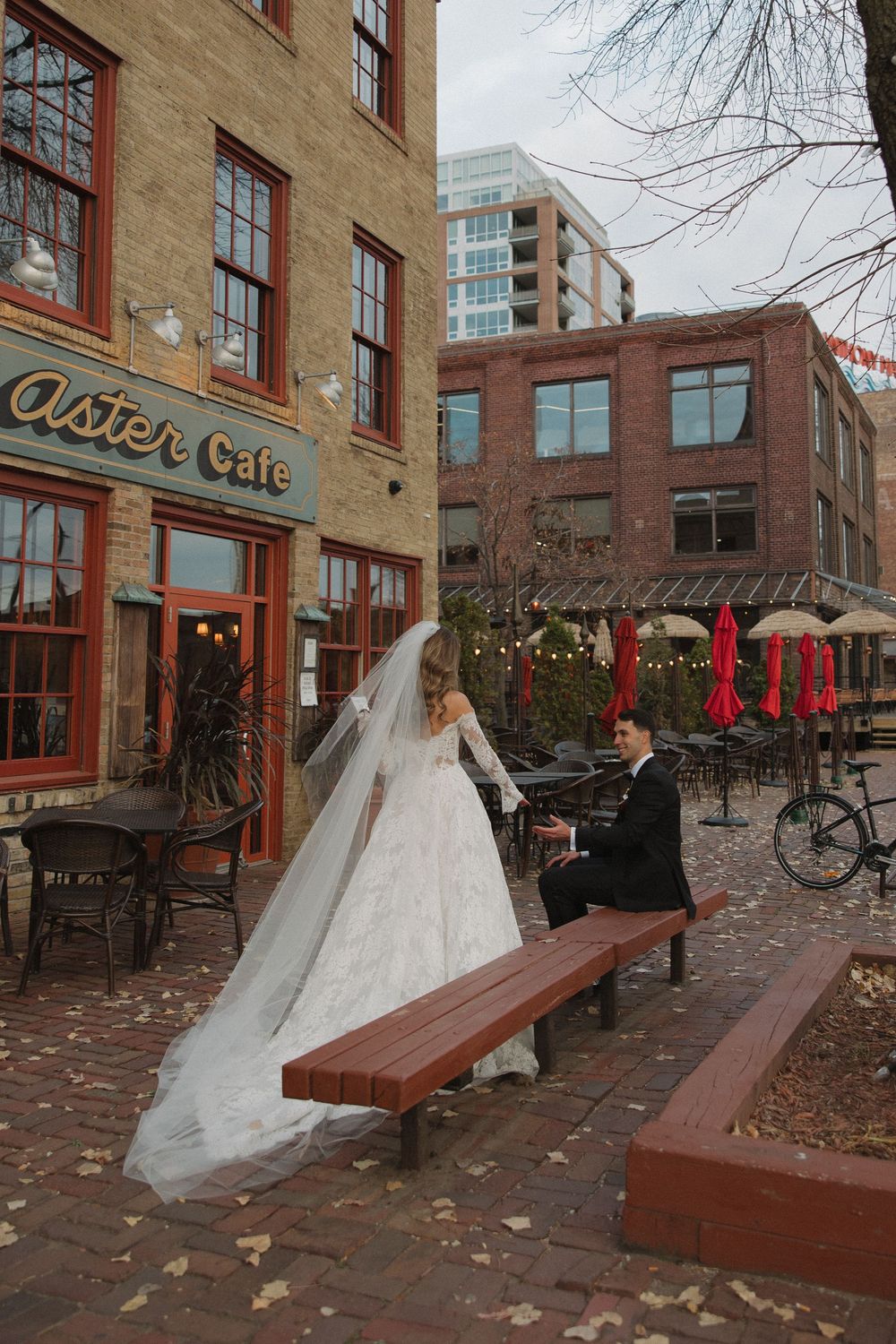 A wedding scene outside a brick building with a cafe, featuring a wooden bench and scattered autumn leaves.