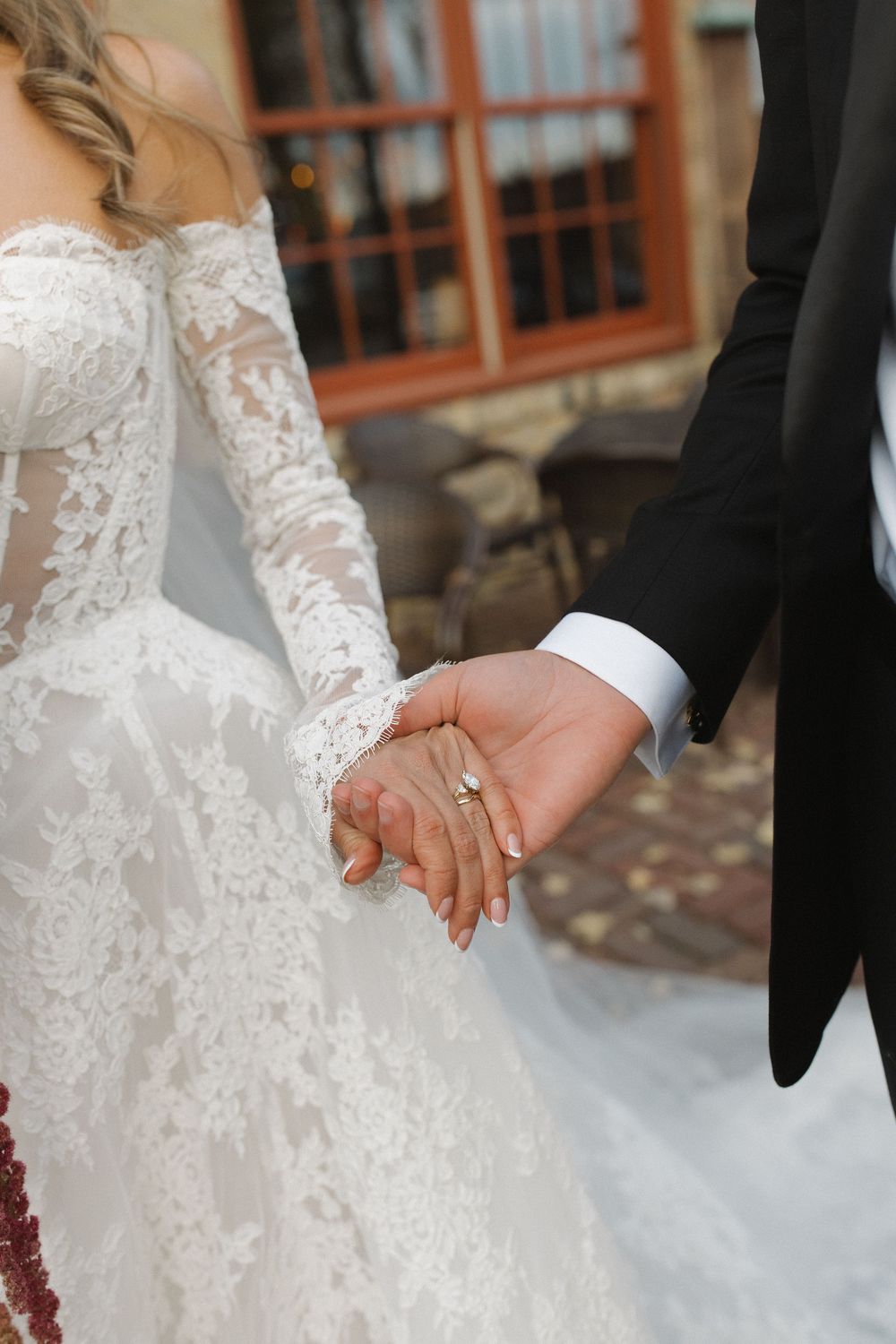 Close-up of newlyweds holding hands showing wedding rings and detailed lace sleeve of white bridal gown.