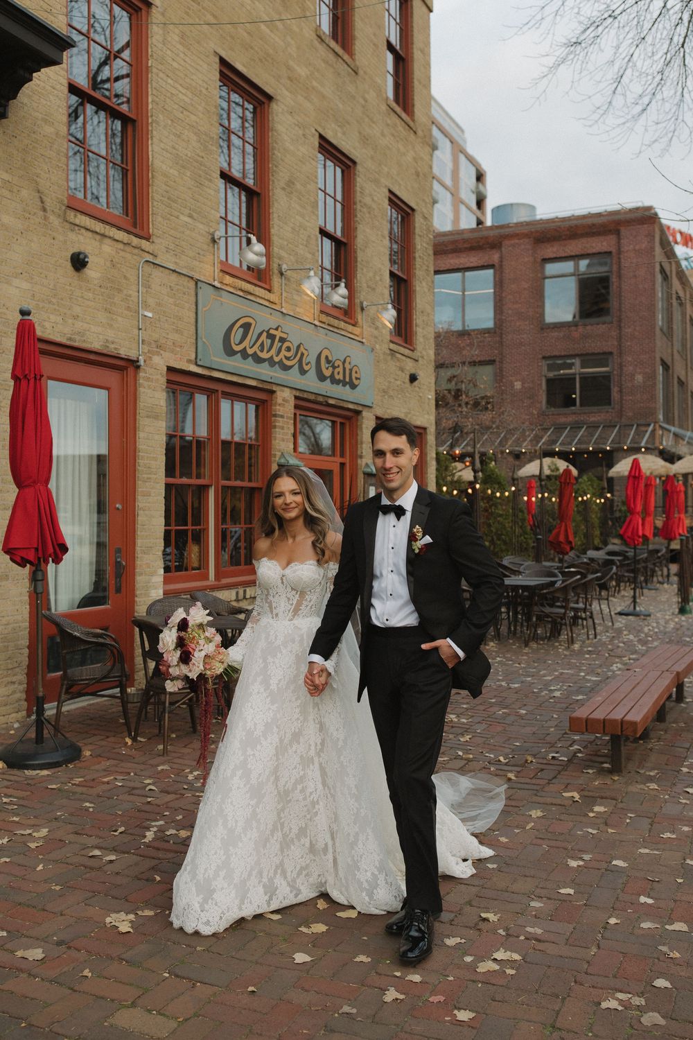 A wedding couple walks along a charming brick street with historic buildings and red umbrellas in the background.