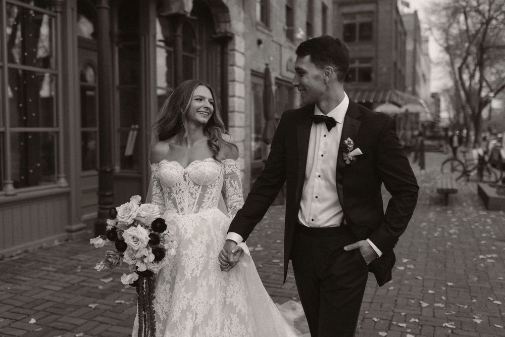 A couple in formal wedding attire walks together on a tree-lined cobblestone street in autumn.