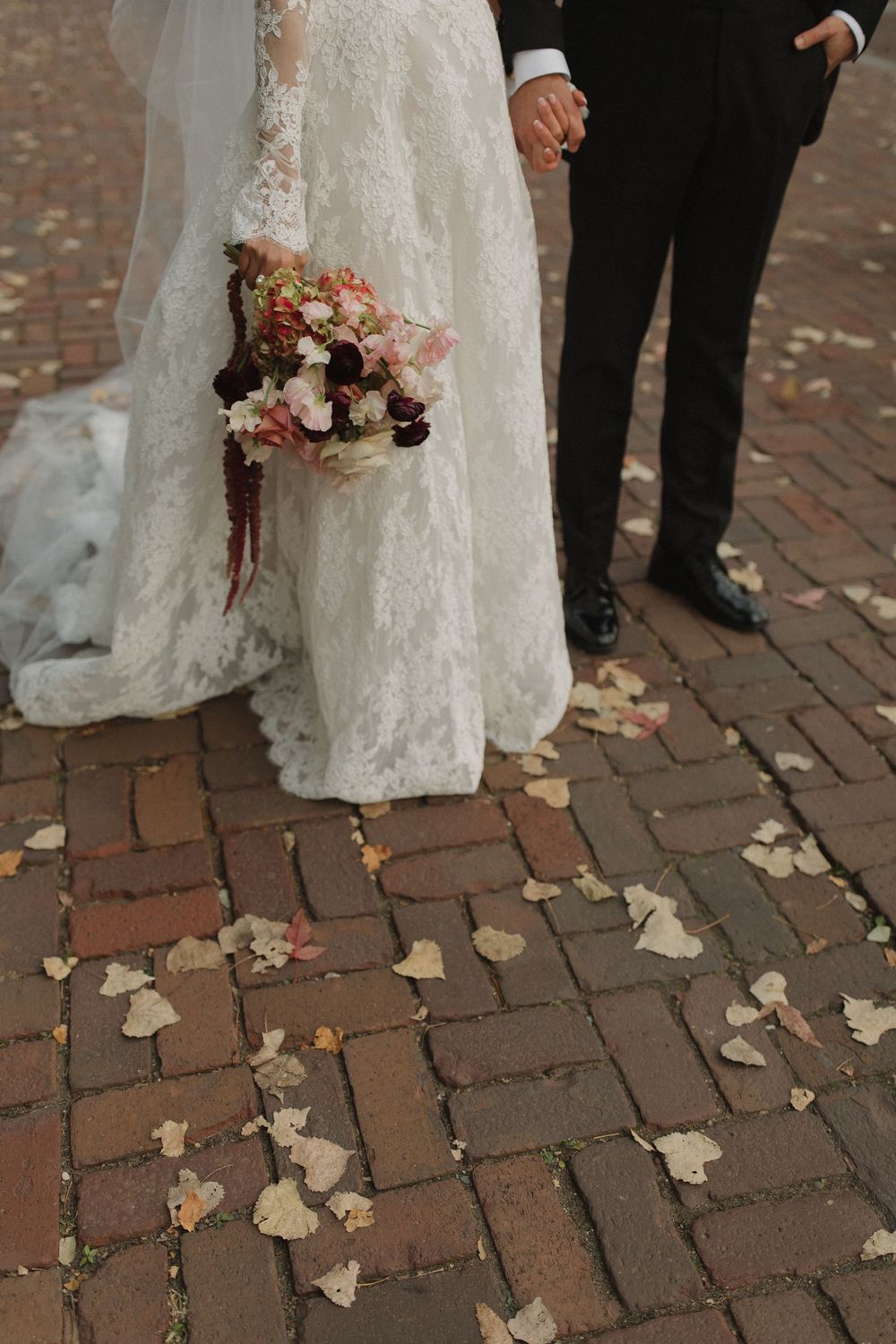 Close-up details of a white lace wedding dress and bouquet against brick pavers covered in fallen leaves.
