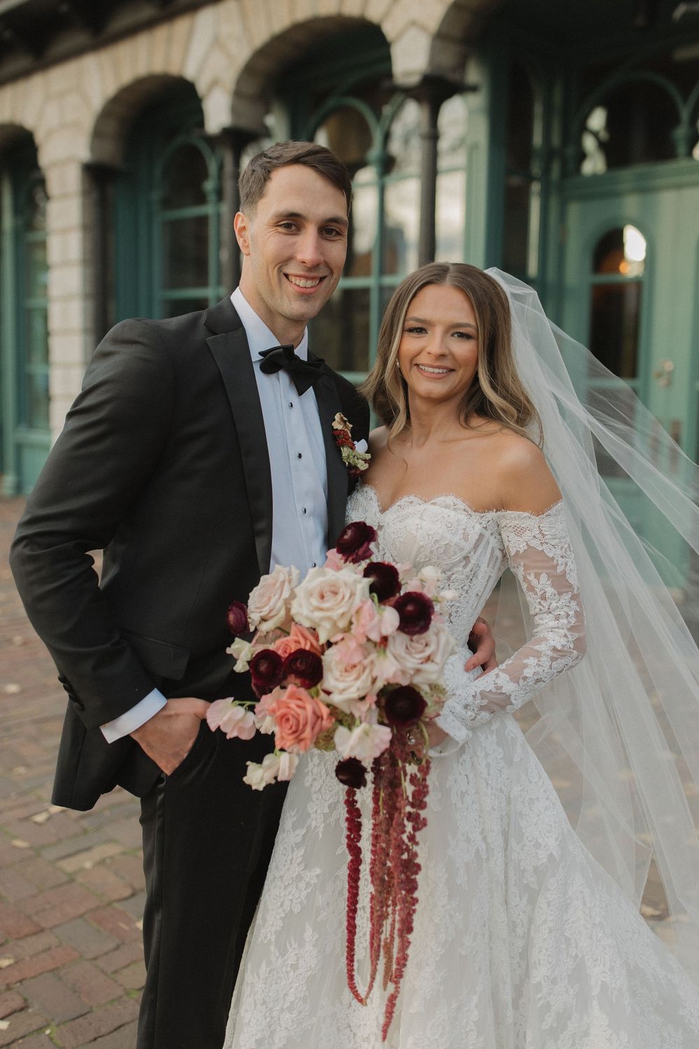 A wedding couple poses in front of a historic building with a stunning bouquet of pink and burgundy flowers.