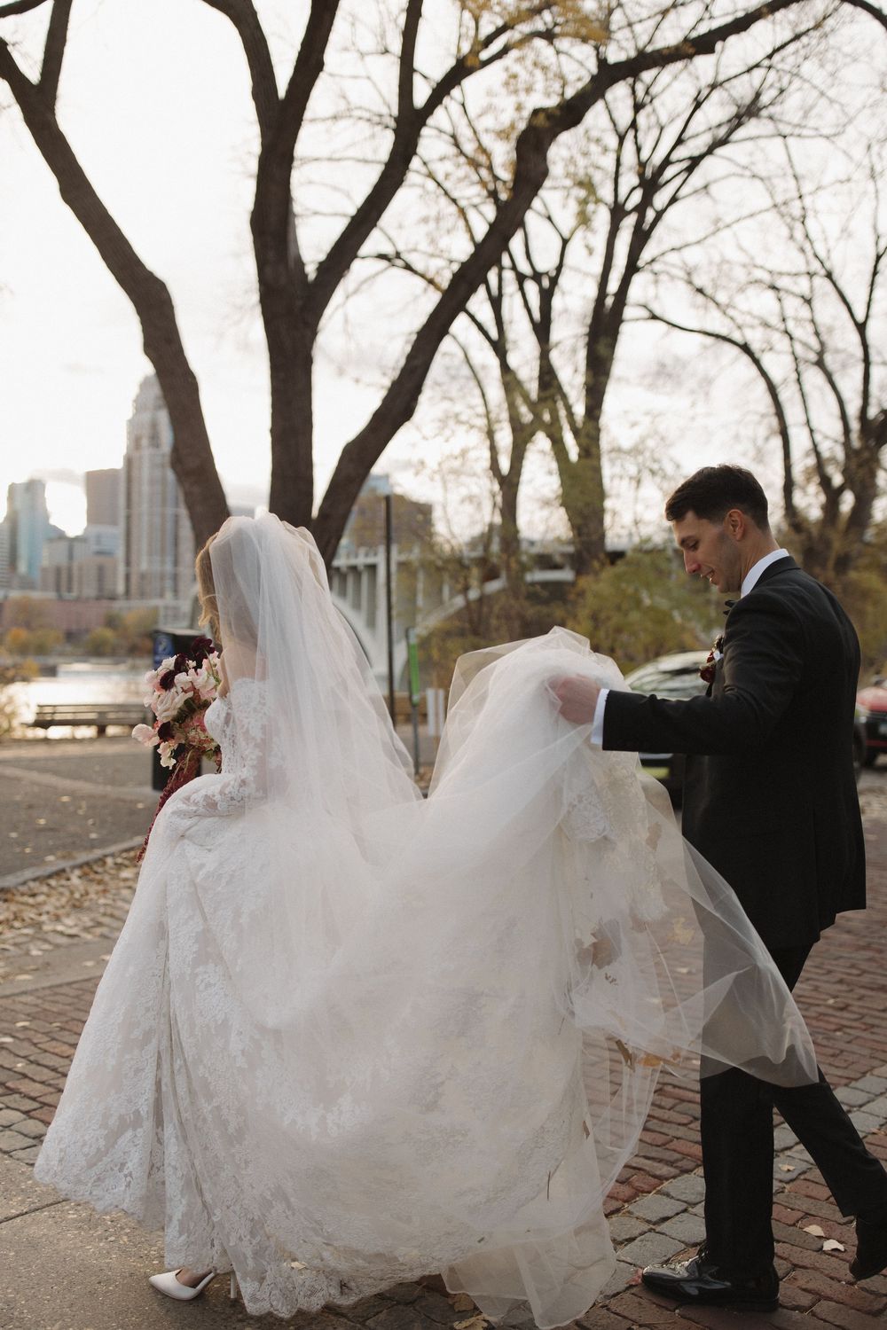 A wedding dress billows in the wind as the couple walks along a city path with autumn trees and skyline.