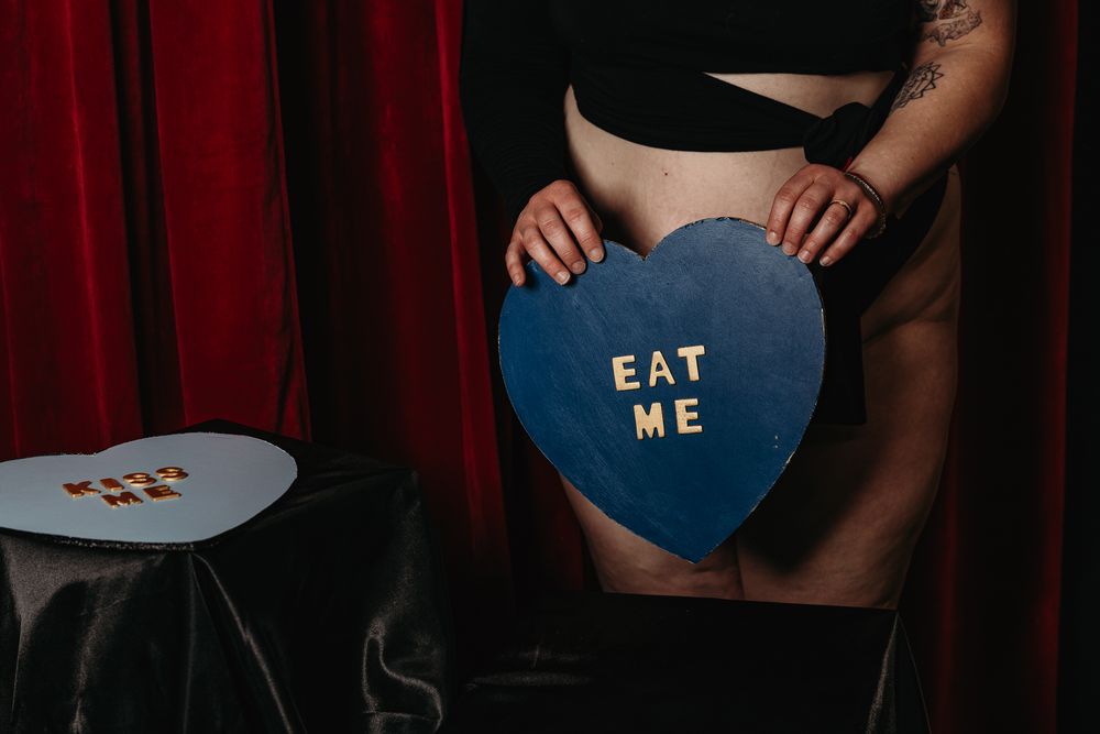 A blue heart-shaped sign with gold text reading 'EAT ME' held against a dark background with red curtains.