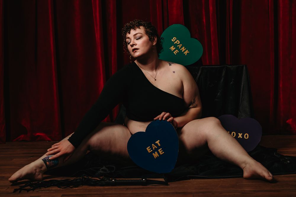 Sensual portrait against deep red curtains with emerald green book creating dramatic contrast.