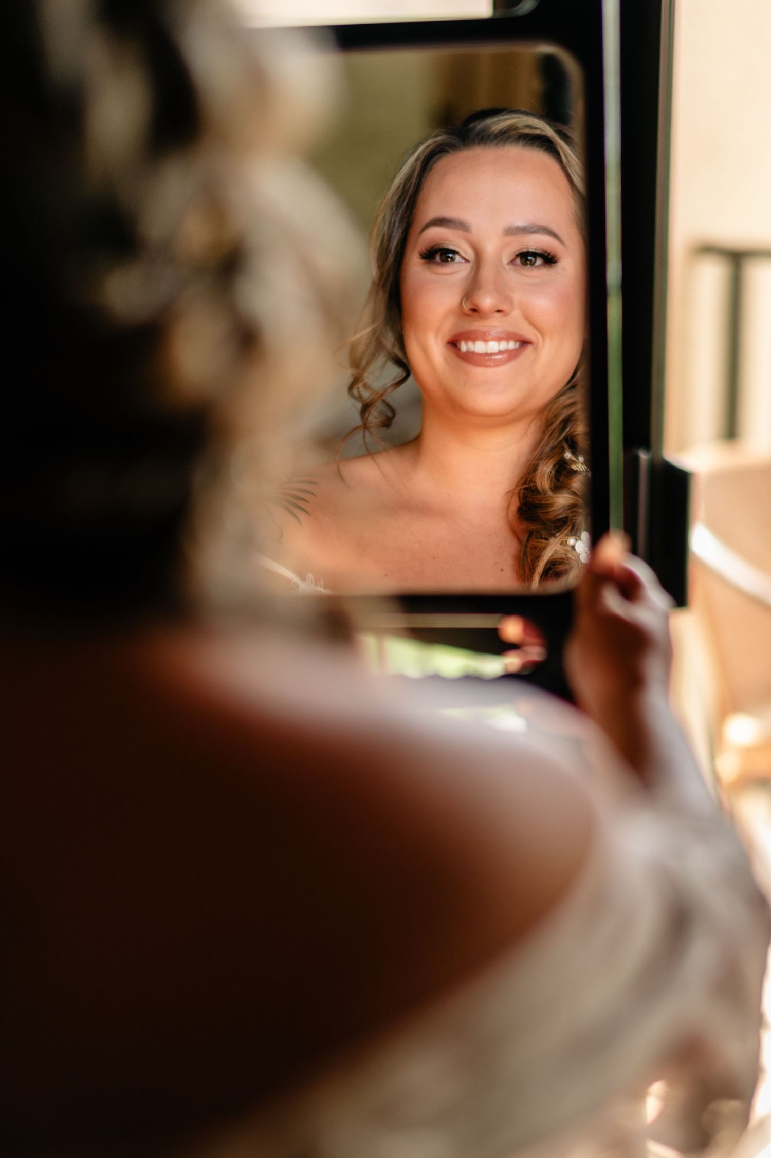 A reflection in a mirror shows a smiling bride with curled hair and dangling earrings on her wedding day.