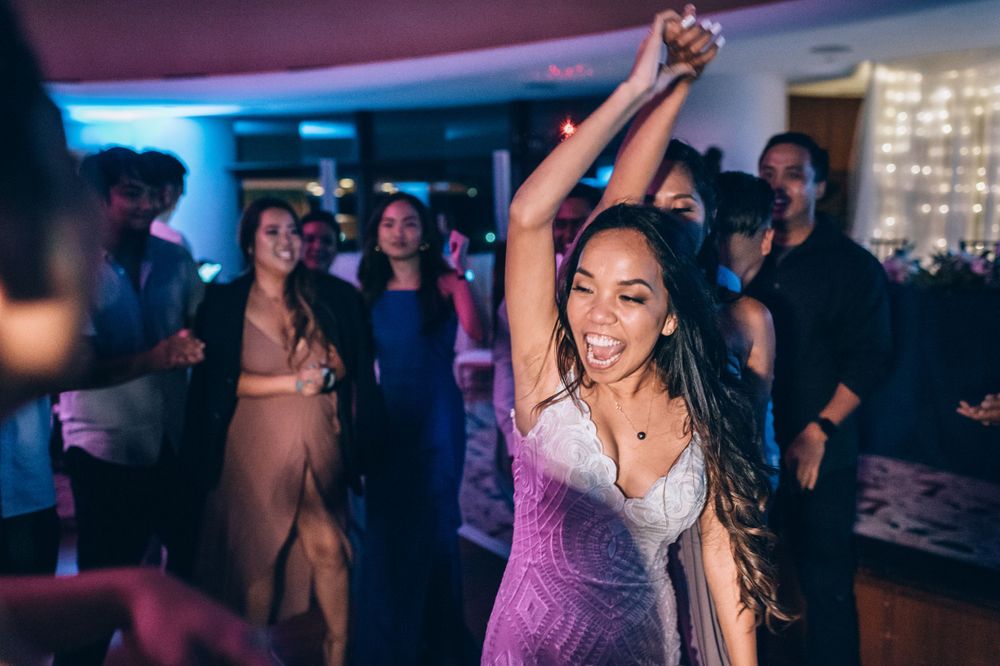 Party guests dancing and celebrating under pink and purple mood lighting at an indoor event.