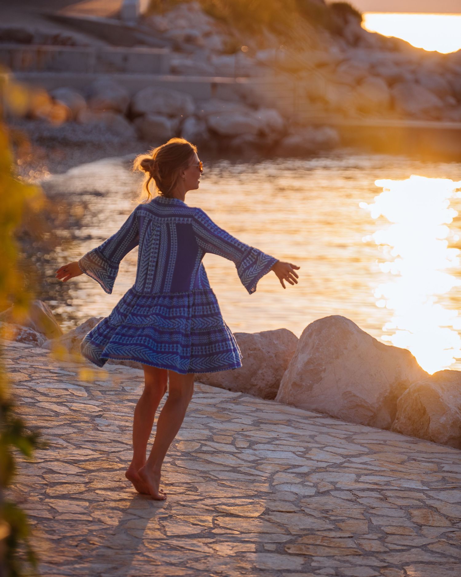 A silhouetted figure in a blue dress twirls barefoot by the water at sunset on a rocky shore.