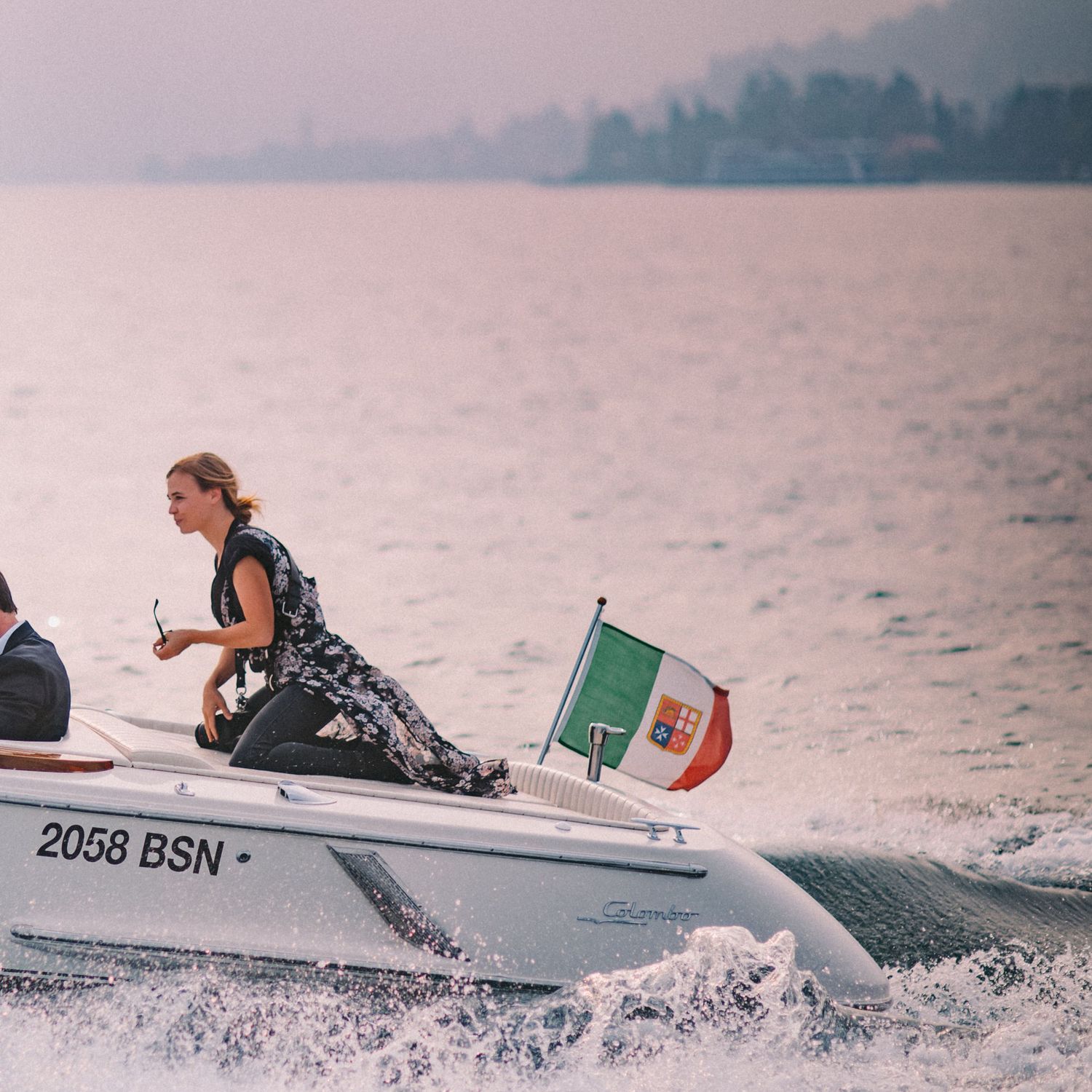 A speedboat cuts through misty waters with an Italian flag visible, creating a dramatic wake on the lake surface.