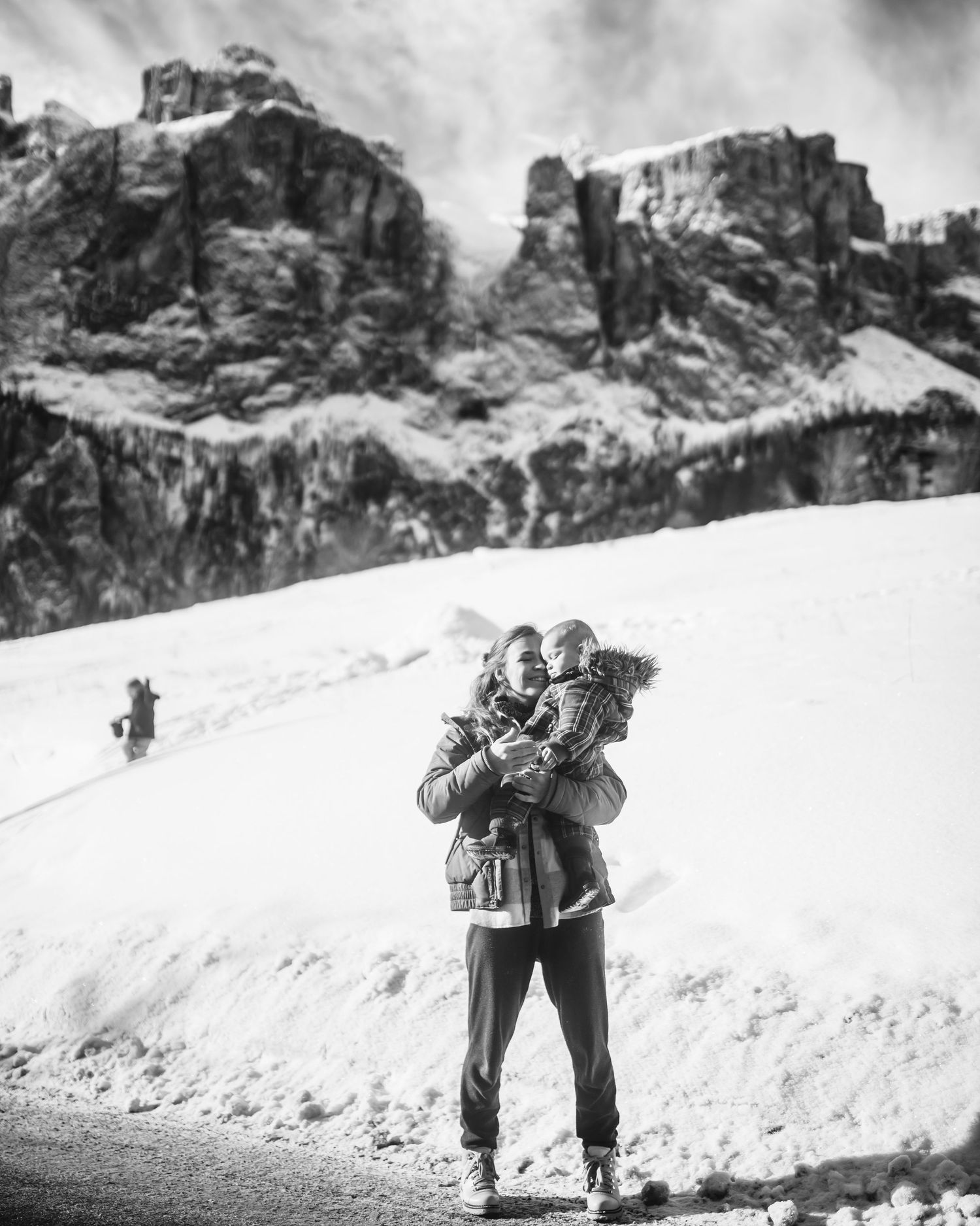 A person wearing winter gear holds a small figure against a snowy mountain backdrop in a black and white photograph.