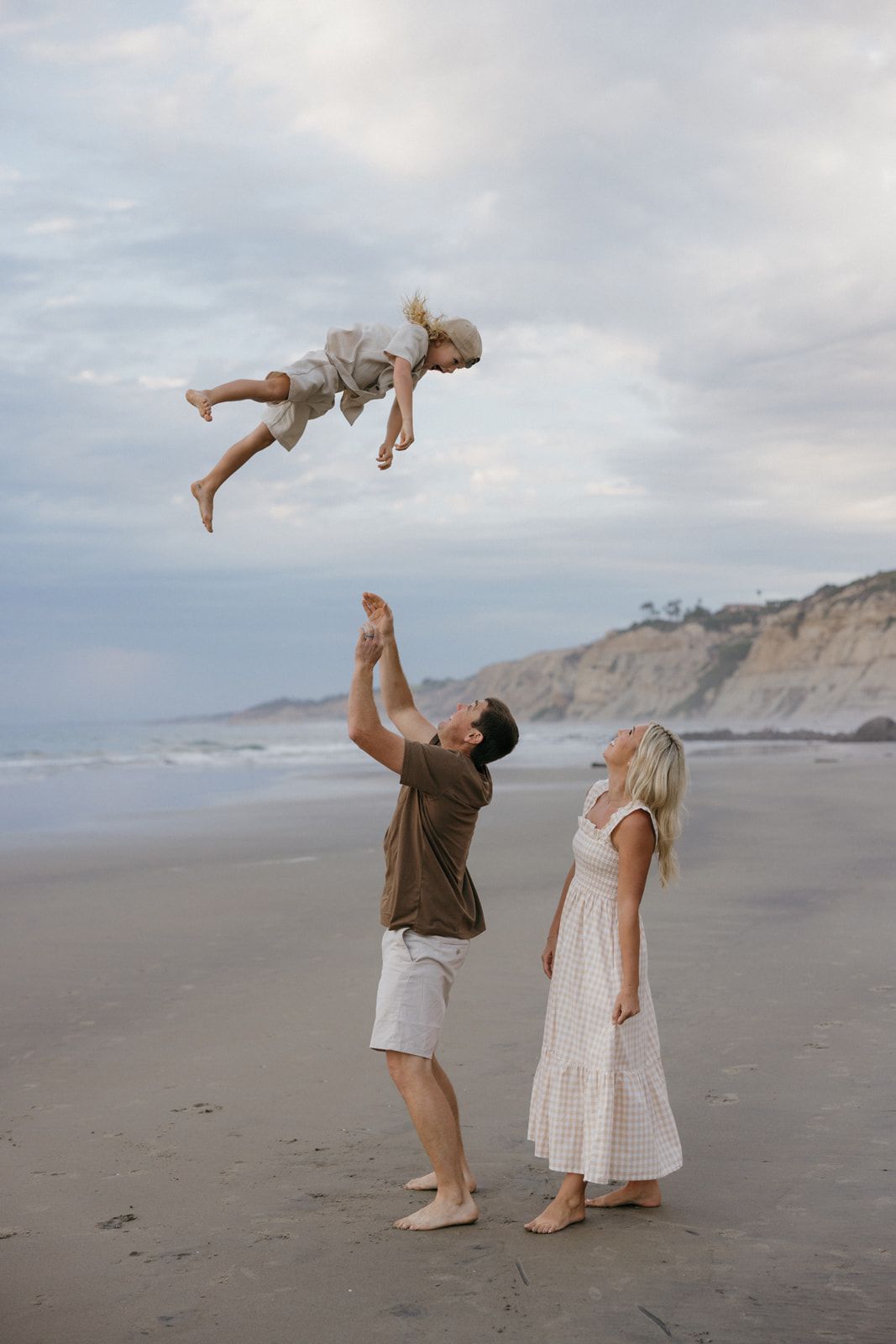 A series of photos showing people playfully tossing a small child in the air at a cloudy beach with cliffs in background.