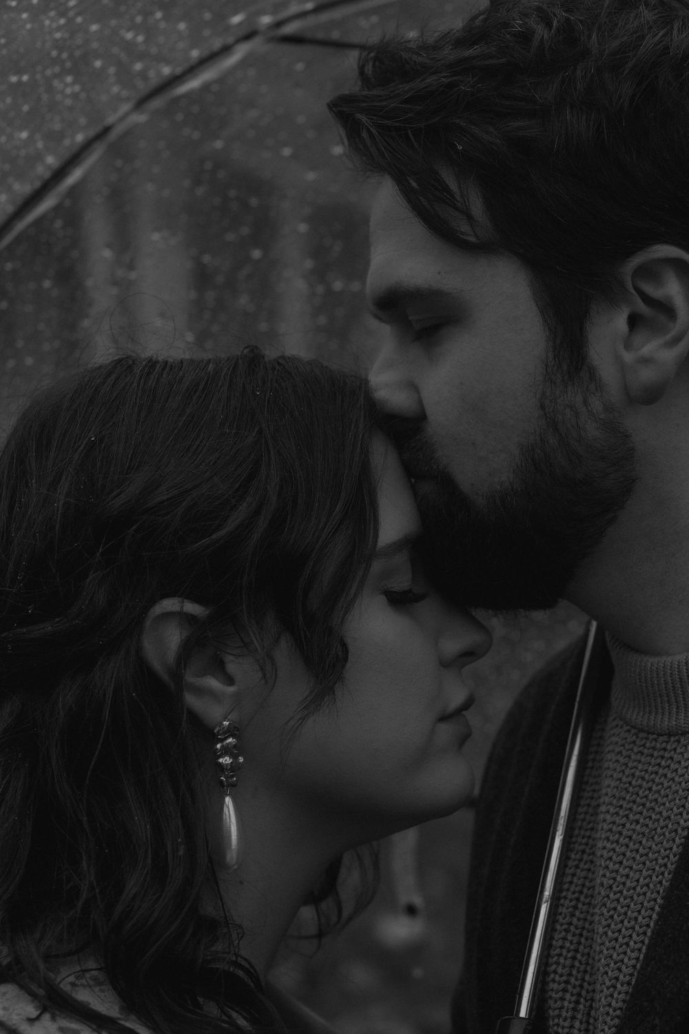 An intimate black and white portrait of a couple in a tender moment against a dark background.