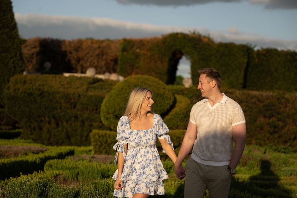 A couple walks hand in hand through a garden at sunset, with manicured hedges and warm golden light.