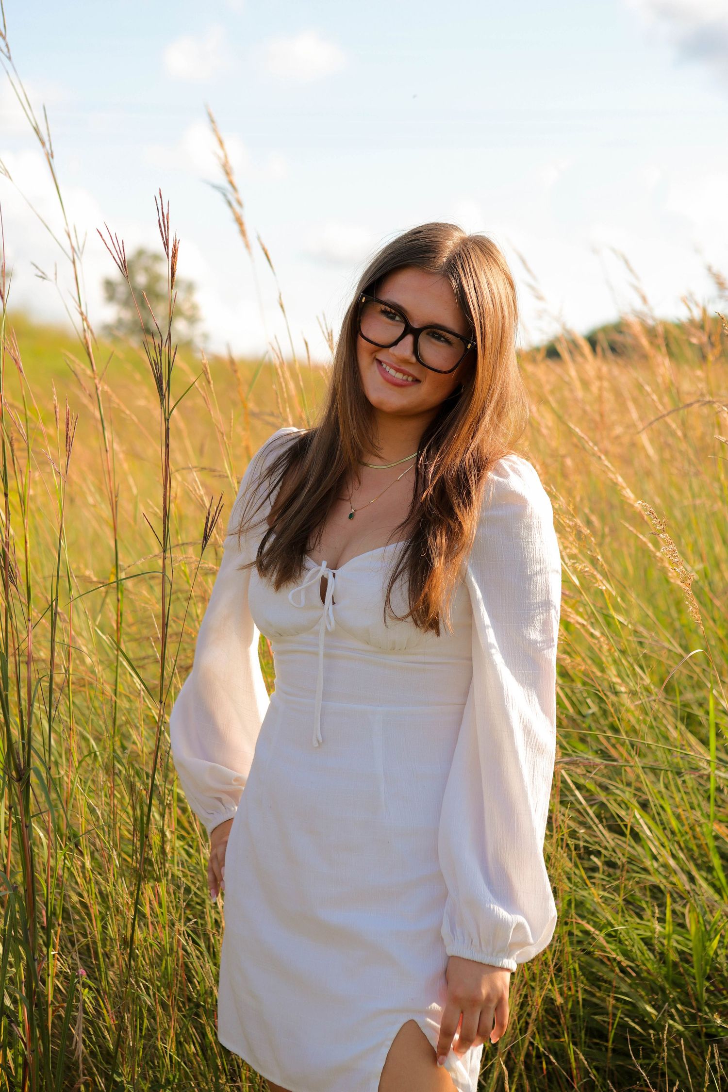 Young woman in white dress and glasses smiling in a sunny golden grass field.