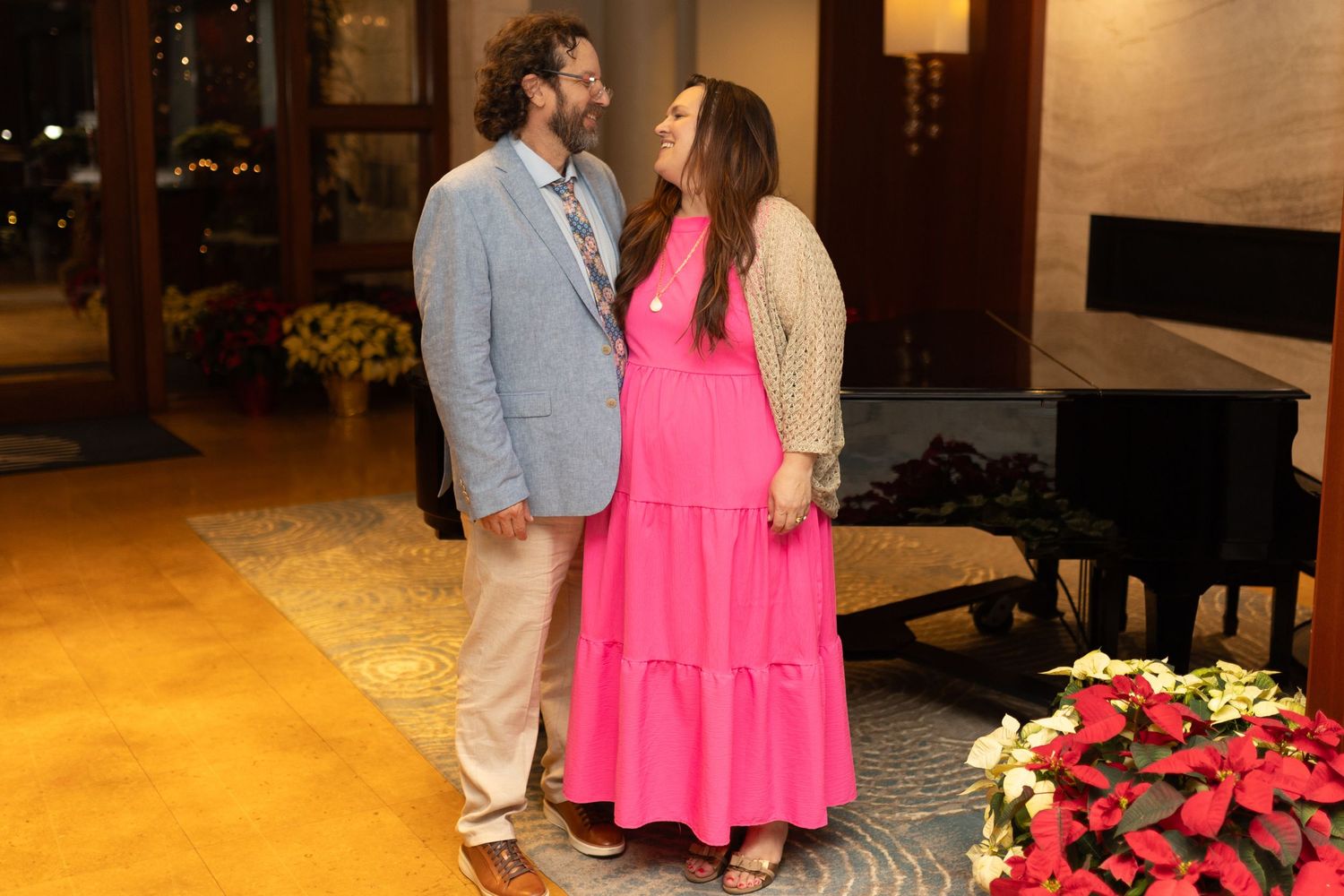 Pregnant woman in pink dress with partner posing in elegant holiday decorated lobby.