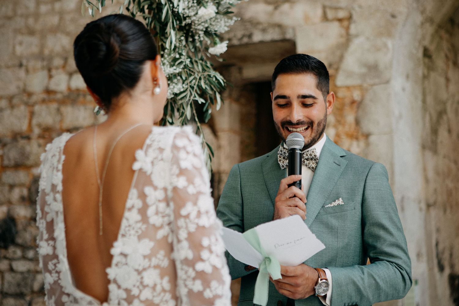Groom in sage green suit reads heartfelt vows into a microphone to his bride during outdoor wedding ceremony.