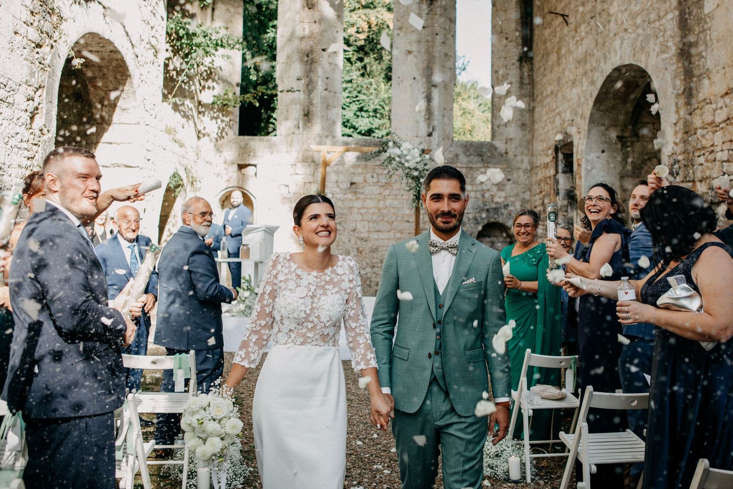Newlyweds walk down the aisle amid confetti celebration surrounded by guests in a beautiful stone ruin venue.
