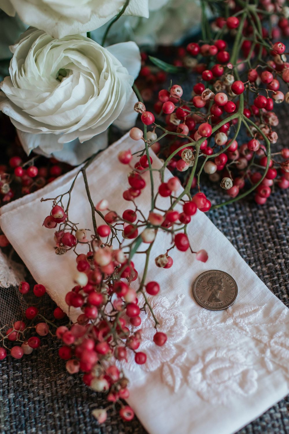 White ranunculus flower with red berries arranged on embroidered fabric with vintage coin detail.