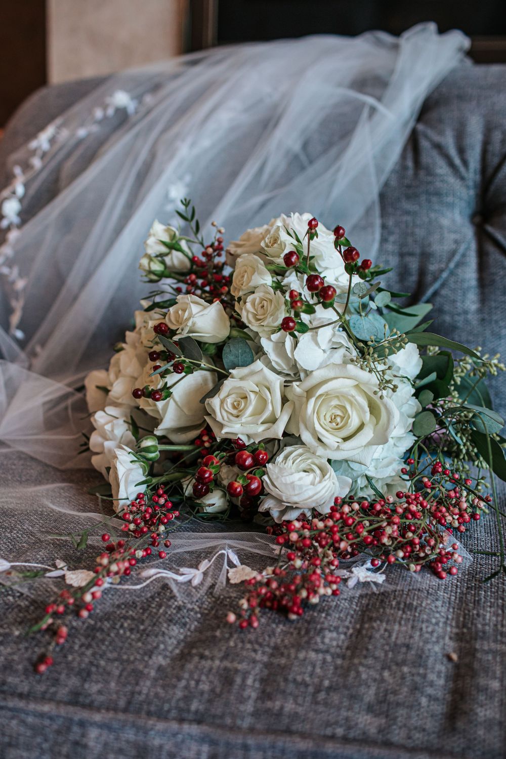 A delicate winter bridal bouquet with white roses, red berries, and eucalyptus leaves rests on gray fabric.
