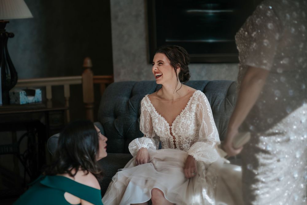 A joyful bride in a lace wedding dress laughs while getting ready with her bridal party.