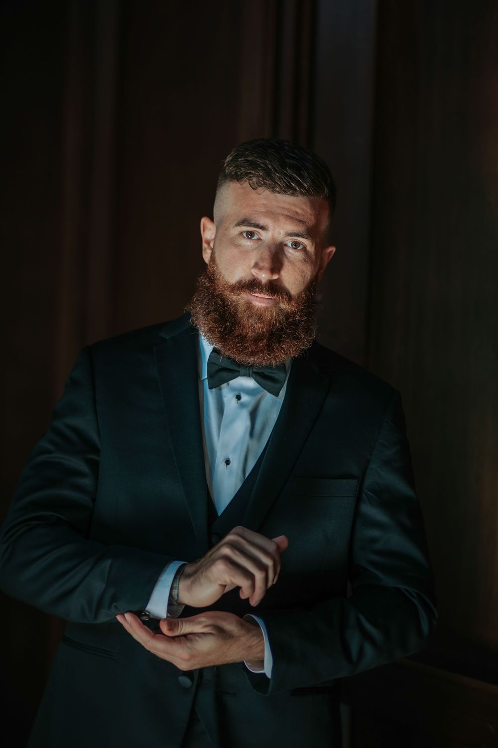 A bearded man in a dark suit and light blue tie poses in dramatic low-key lighting against a black background.