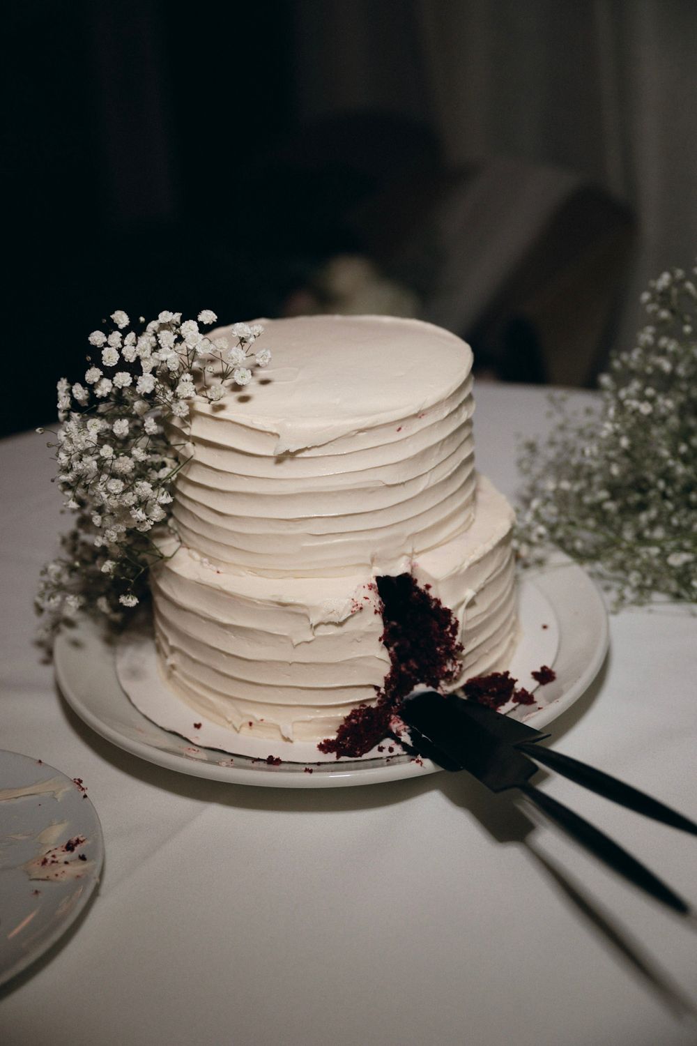 Elegant white wedding cake with textured frosting and baby's breath flowers being cut on a white tablecloth.