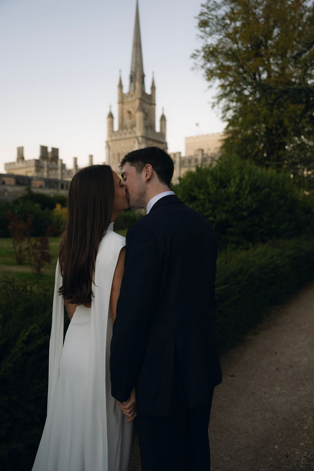 A couple in wedding attire share a romantic moment with a historic church spire visible in the background.